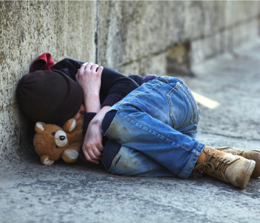 A child in an alley curled up with a teddy bear